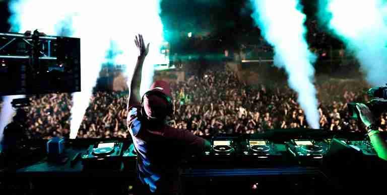 A DJ performs on stage, raising one hand toward a large crowd. Smoke effects and colorful lighting enhance the lively atmosphere, making it a perfect scene for captivating social media design.