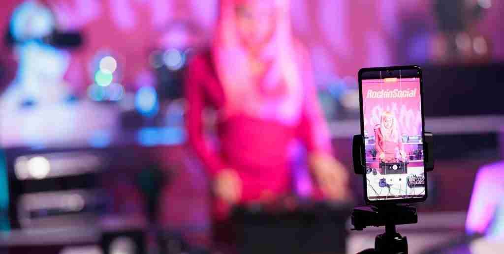 A smartphone on a tripod records a person in a pink hijab standing and speaking, with a blurred audience and pink lighting in the background, perfectly capturing the essence of social media design.
