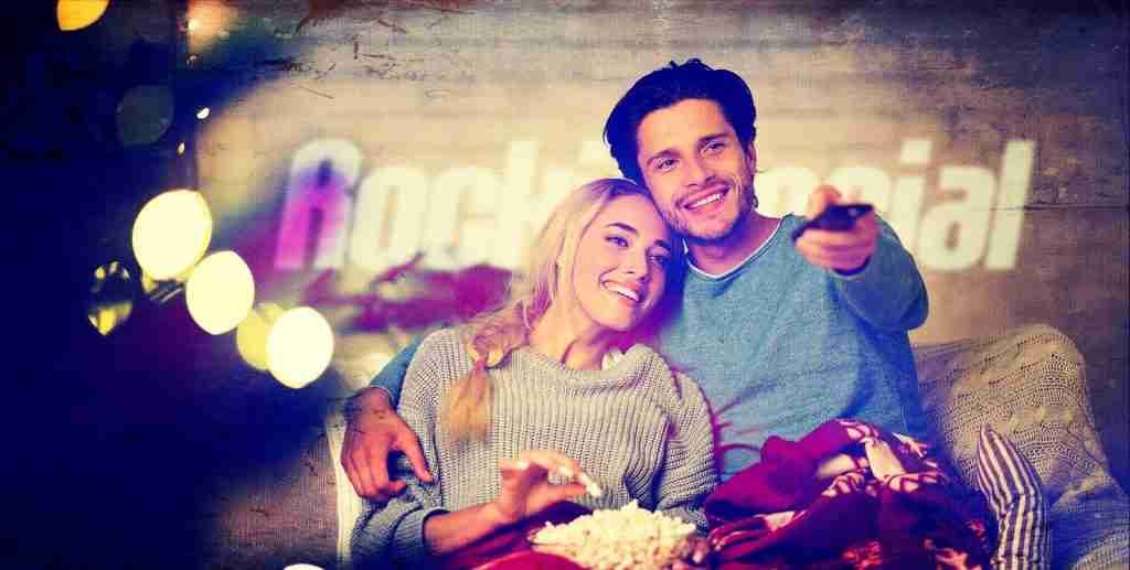 A young couple sits on a couch, smiling and watching TV, with the man holding a remote and a bowl of popcorn on the woman's lap. The background features stylized text, string lights, and hints of social media design elements subtly enhancing the cozy scene.