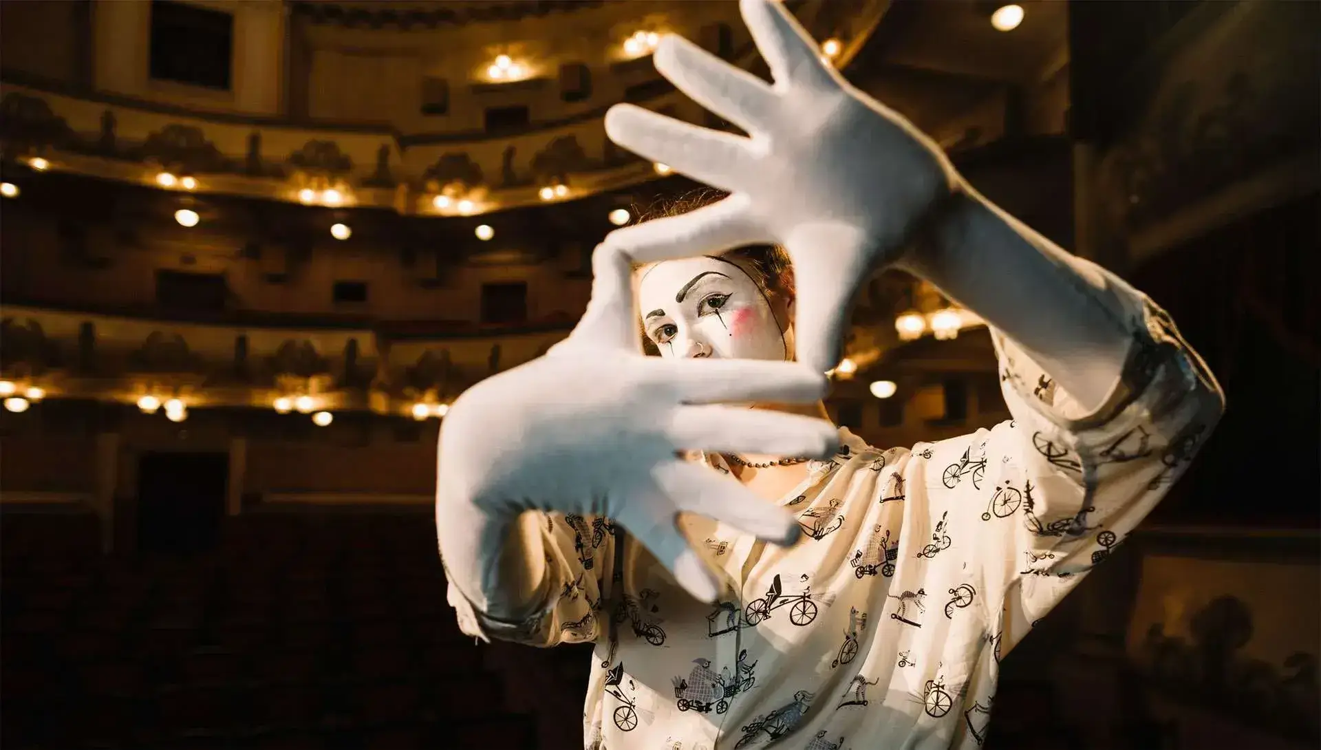 A performer in white gloves and face paint poses dramatically with hands framing their face in an empty, dimly lit theater, creating a scene that would captivate any social media design.